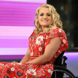 Ali Stroker sits on stage in a wheelchair smiling wearing a pink and red flroral dress