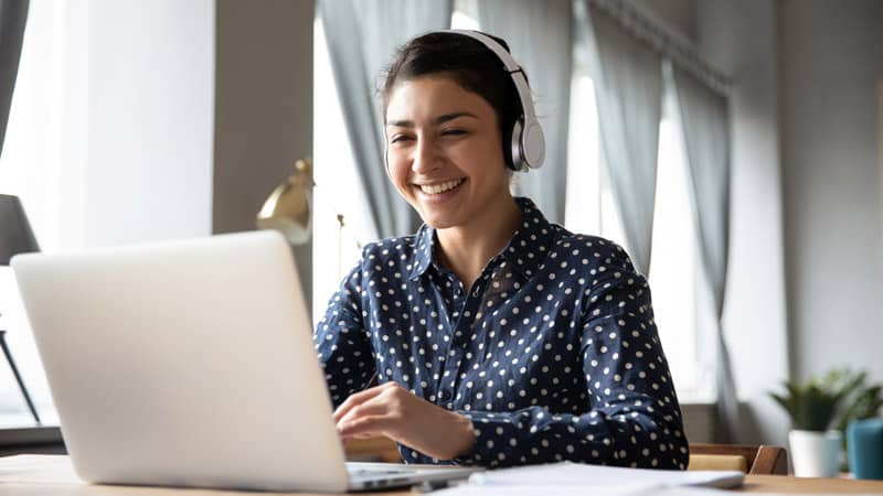 Smiling Indian girl wearing headphones using laptop, networking online