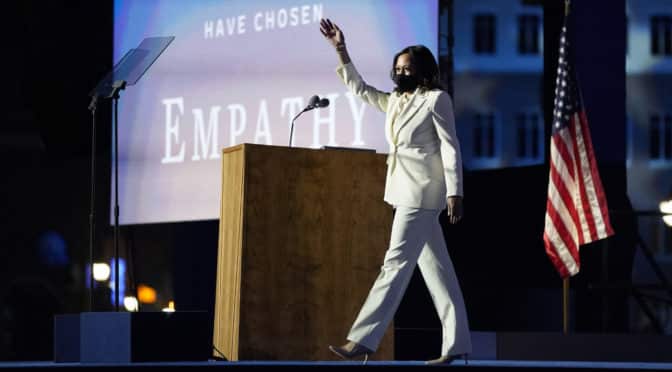 Kamala Harris on stage white suit wearing a mask with flag in background