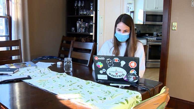 woman sitting at dining table sewing on a sewing machine wearing a mask