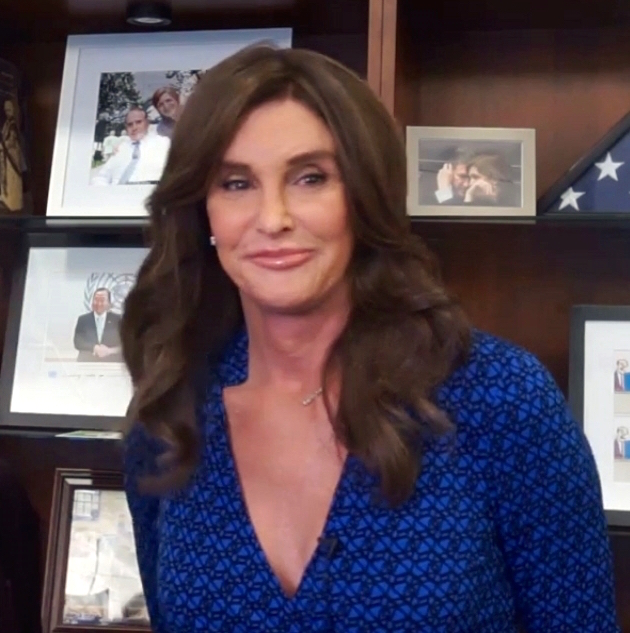 Cailtlyn Jenner in blue dress smiling in front of bookshelf with books and photos