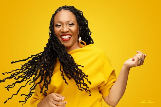 Tara Darnley, founder of Peculiar Roots, posing in front of yellow background in a yellow dress