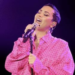 Demi Lovato performs during a premiere event for their documentary wearing an all pink button up in front of a purple background