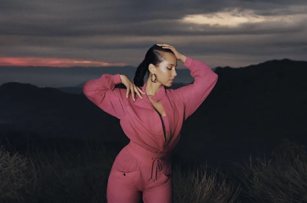 Alicia Keys wearing a pink jumpsuit during sunset posed to the side with one hand over her head and one on her own shoulder