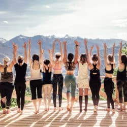Rear view of group of joyful happy athletic young women in gymnastic clothes raise their hands up and enjoy joint activities in open air among mountains. Concept of sports and personal