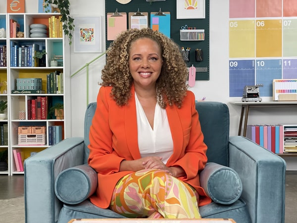 Belva Anakwenze wearing bright orange jacket sitting in large chair smiling